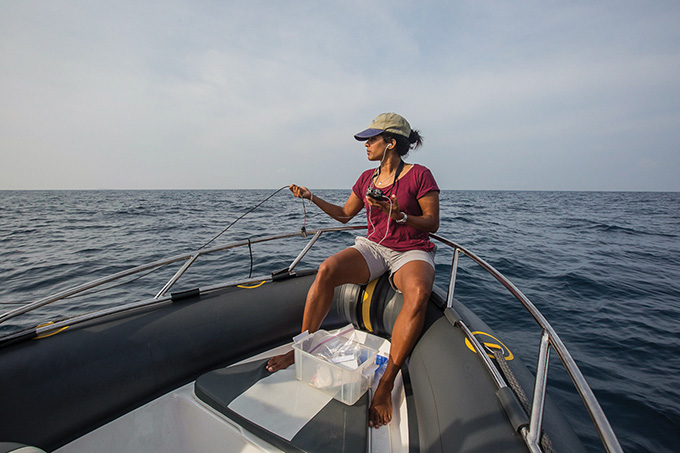 Asha de Vos is sitting on a boat, holding lab equipment in both hands. She is wearing a baseball cap, a maroon colored t-shirt and a white pair of shorts. She is not wearing shoes.